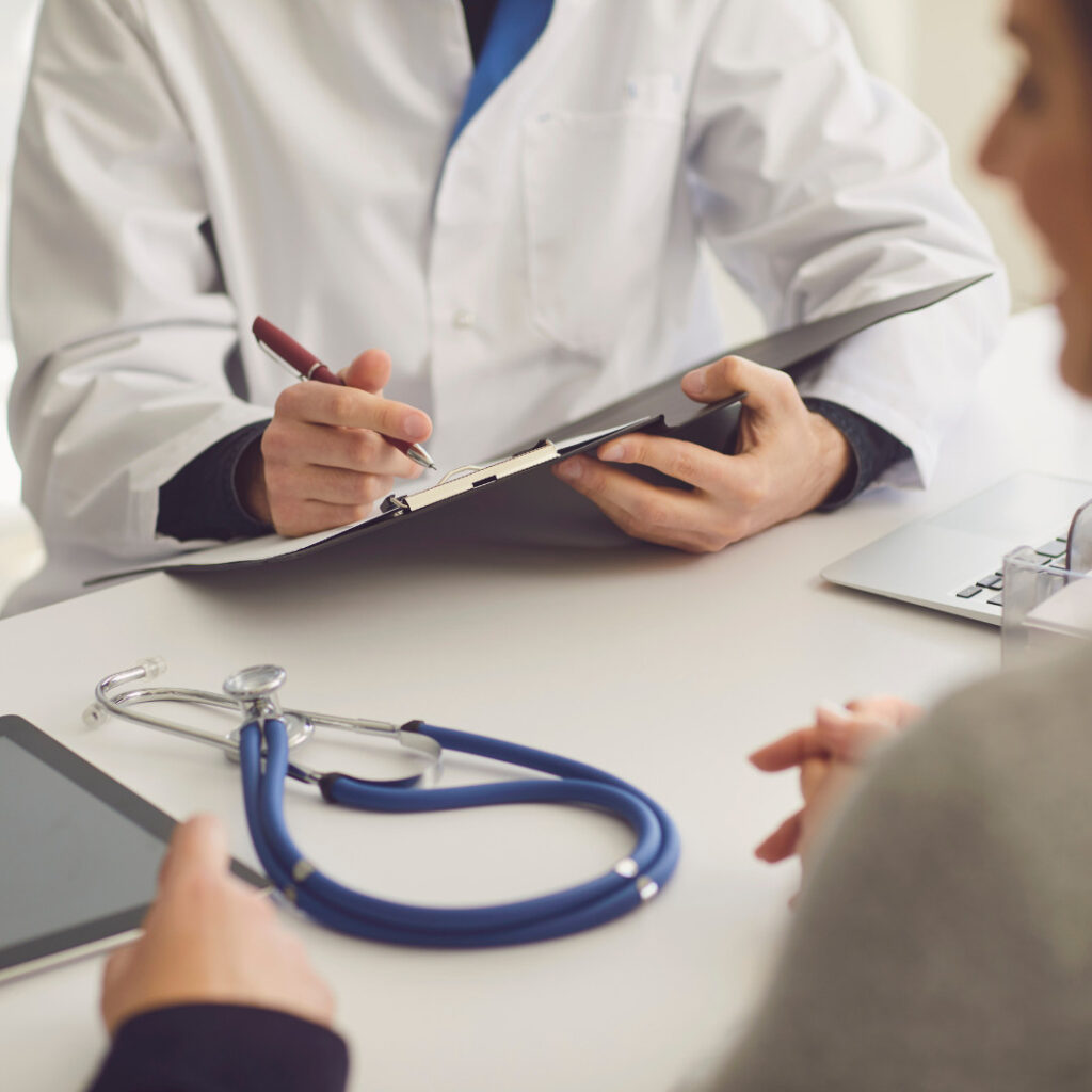 The image shows a doctor wearing a white lab coat, holding a clipboard and pen. A stethoscope is placed on the table nearby. In the foreground, a patient is visible, suggesting a consultation.
