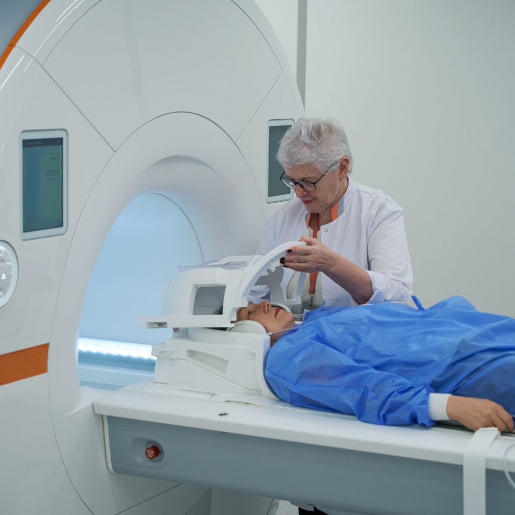 The image shows a medical setting where a healthcare professional is preparing a patient for an MRI scan. The patient is lying down on the MRI machine table, wearing a blue gown. The healthcare worker, wearing a white coat, is adjusting the head coil on the patient.