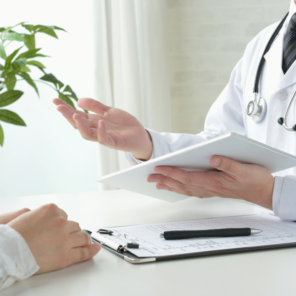 The image features a doctor wearing a white coat and a stethoscope around their neck, holding a tablet while engaged in conversation with a patient. The patient’s hands are visible on the table, where a survey with a pen and paper is also present.