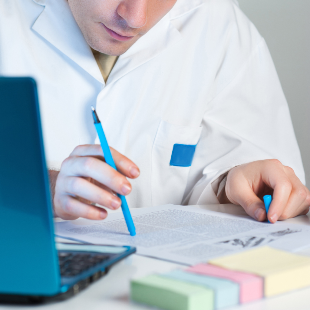 The image shows a scientist reviewing scientific research with a blue pen in hand. Pastel-colored sticky notes help organize information. An open laptop suggests the use of digital tools alongside printed materials.