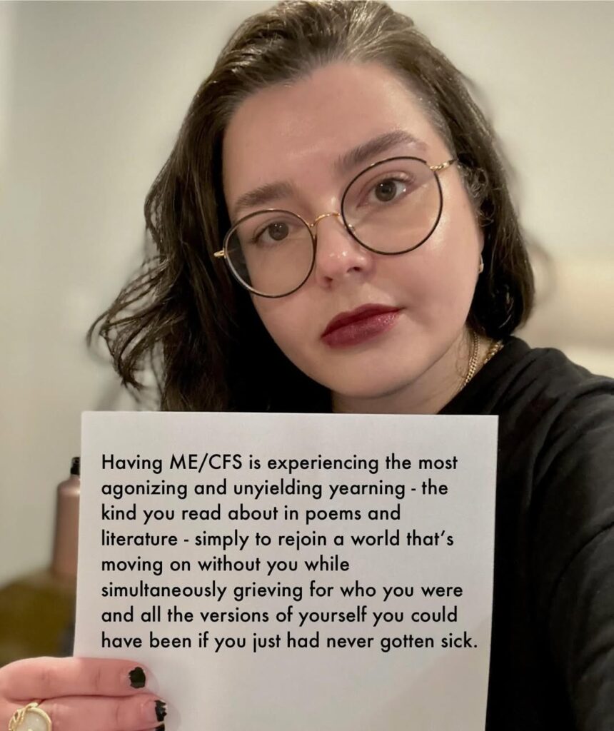 Olenka is holding a sign that reads: Having ME/CFS is experiencing the most agonizing and unyielding yearning-the kind you read about in poems and literature-simply to rejoin a world that’s moving on without you while simultaneously grieving for who you were and all the versions of youself you could have been of you just had never gotten sick.