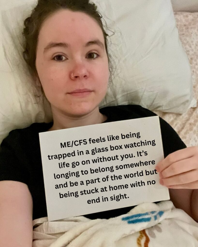 Hayden is holding a sign that reads: “ME/CFS feels like being trapped in a glass box watching life go on without you. It’s longing to belong somewhere and be a part of the world but being stuck at home with no end in sight.