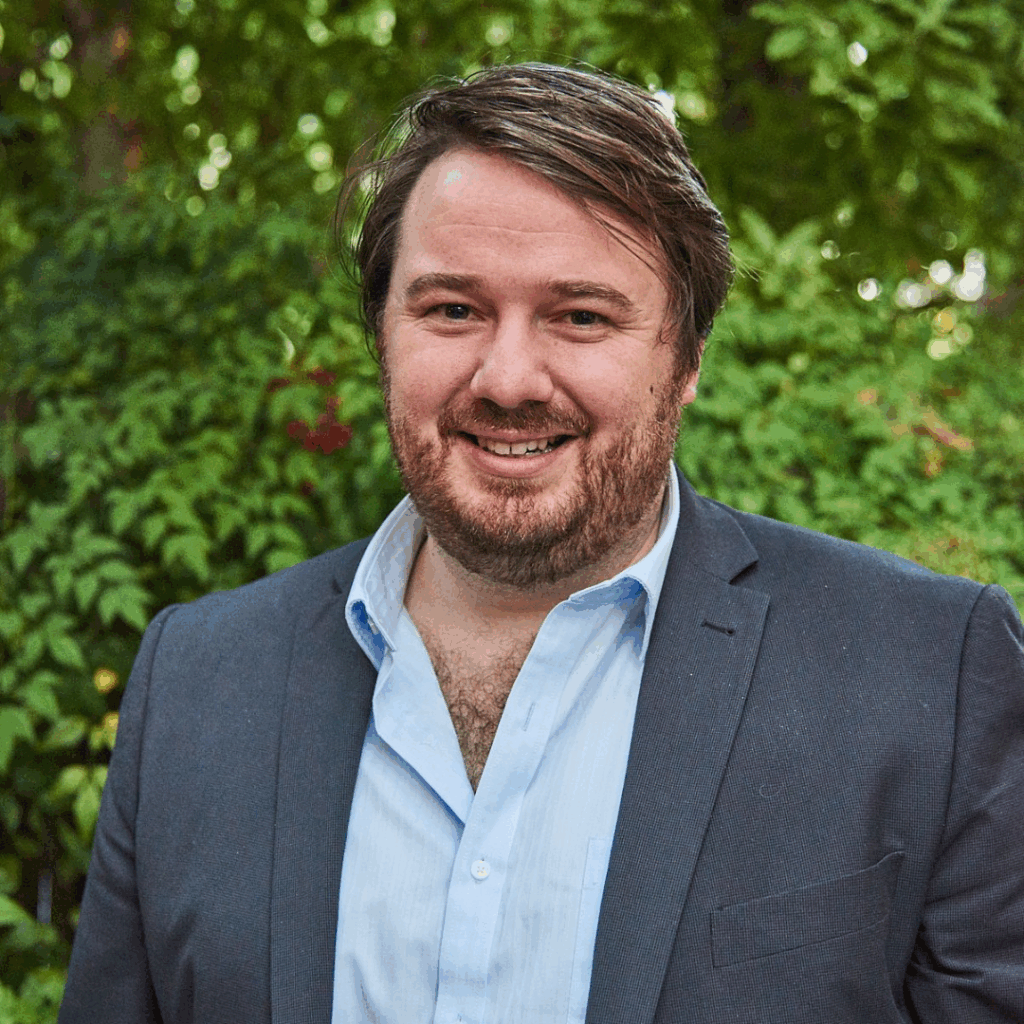 The image shows Dr. Chris Armstrong standing outdoors with greenery in the background. He has short brown hair, and a beard. He is smiling and wearing a light blue, partially unbuttoned dress shirt under a dark grey blazer.