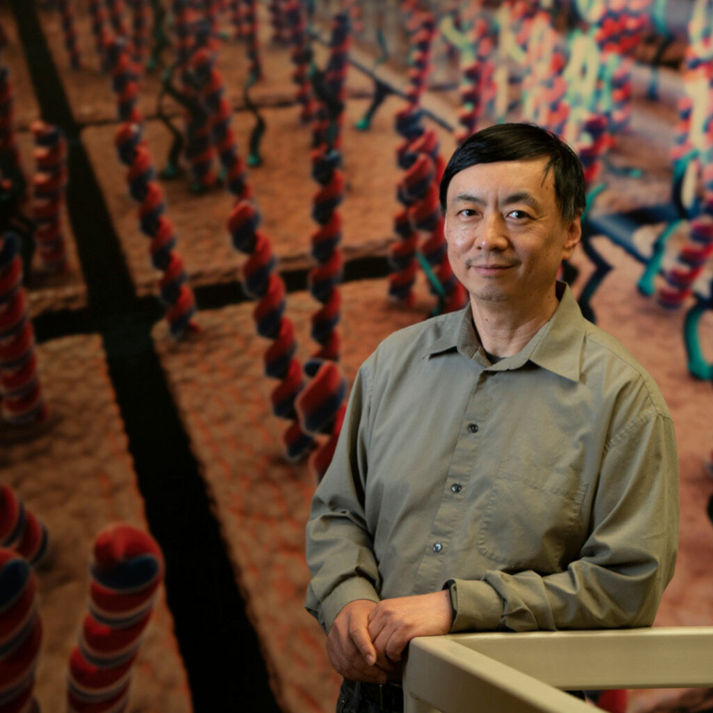 The image shows Wenzhong Xiao standing indoors, leaning lightly on a railing. Behind him is a large, colorful scientific display featuring oversized, twisted, rope-like structures in red and blue, arranged in repeating rows.