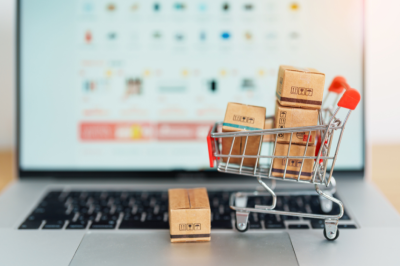 The image shows a miniature shopping cart filled with small cardboard boxes, placed on a laptop keyboard. In the background, the laptop screen displays a blurred online shopping site.