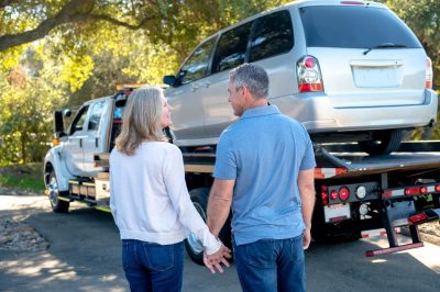 The image depicts a couple holding hands and standing in front of a tow truck that has a silver SUV loaded onto it. They are outdoors, with trees and sunlight in the background.